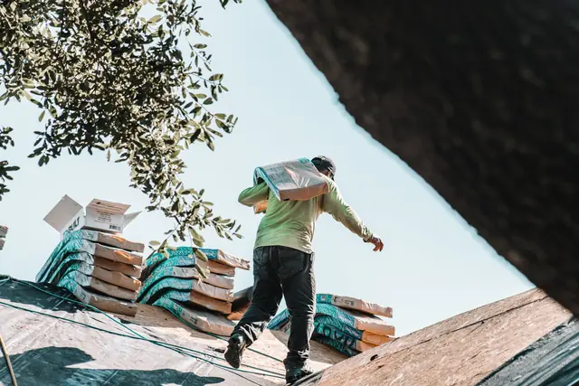 A roofer carries a bundle of shingles across a house roof